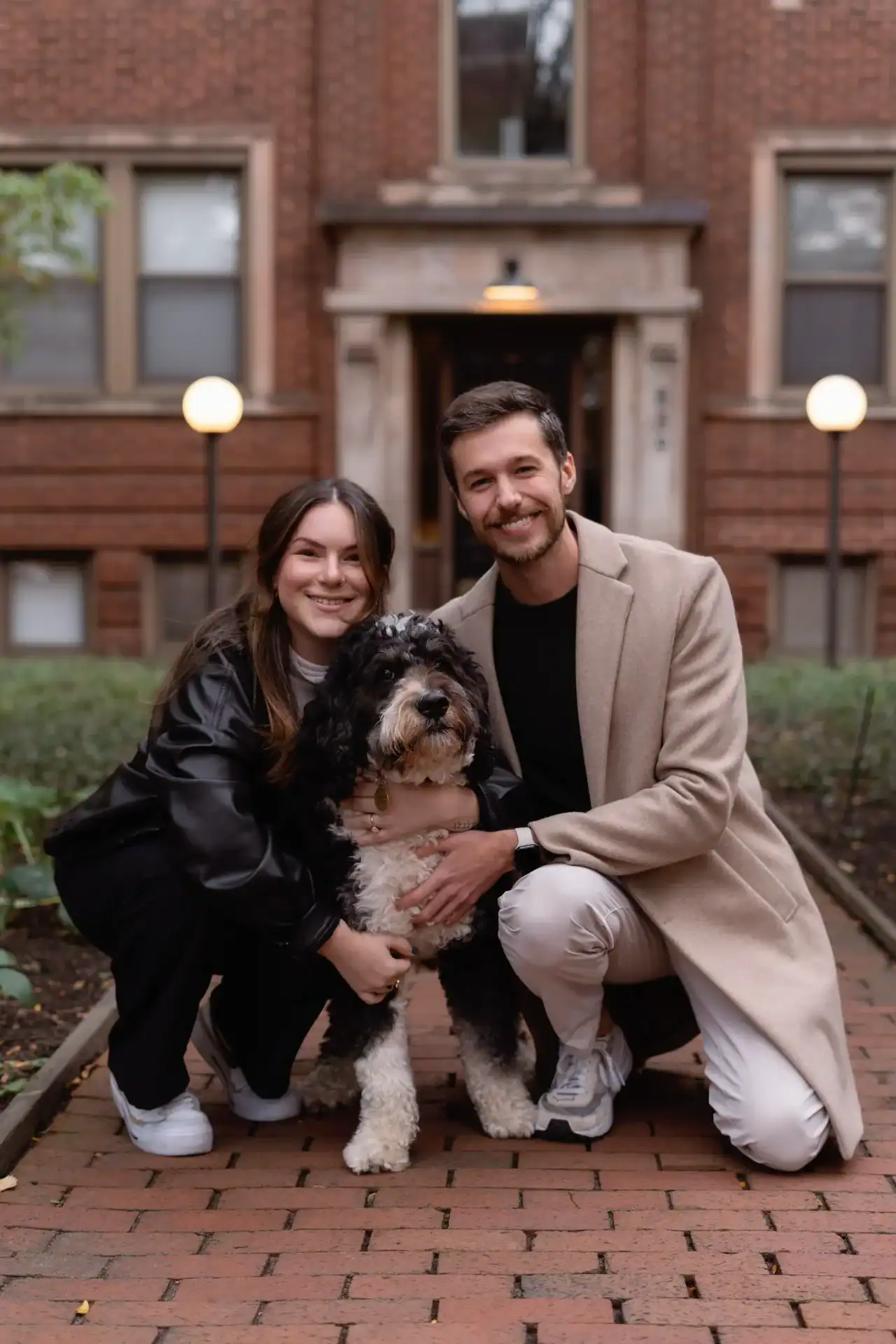 Husband and wife posing with their Bernedoodle on a brick sidewalk in the fall